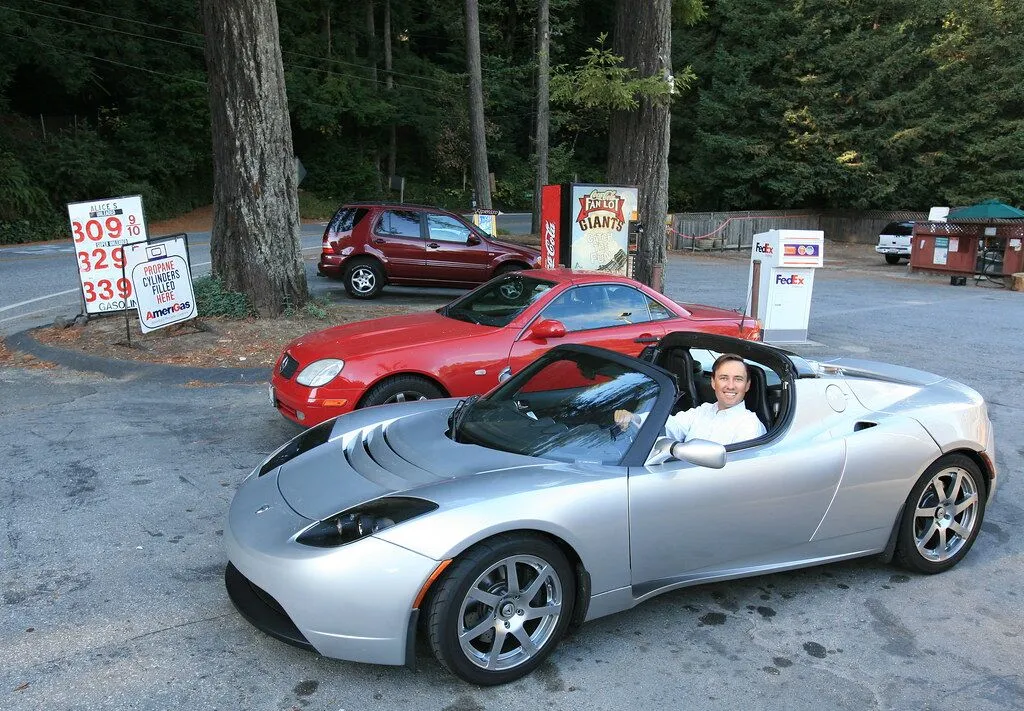 Silver sports car at gas station with red vehicle and trees in background